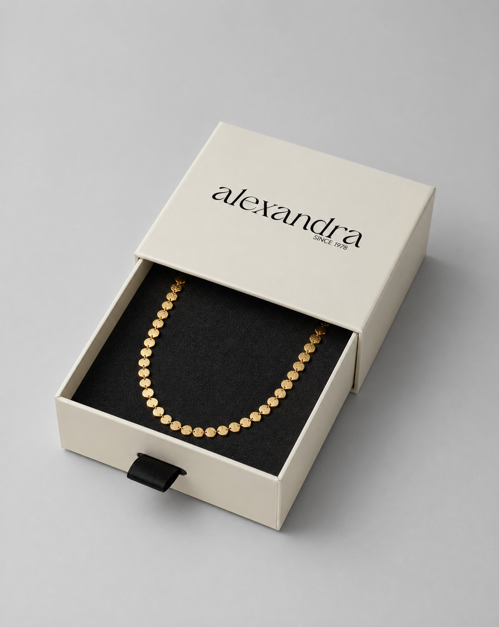 Delicate gold necklace in an open 'alexandra' jewelry box on a gray background