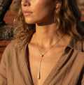 Close-up of a woman wearing a gold lariat necklace with a pendant, set against a natural background.