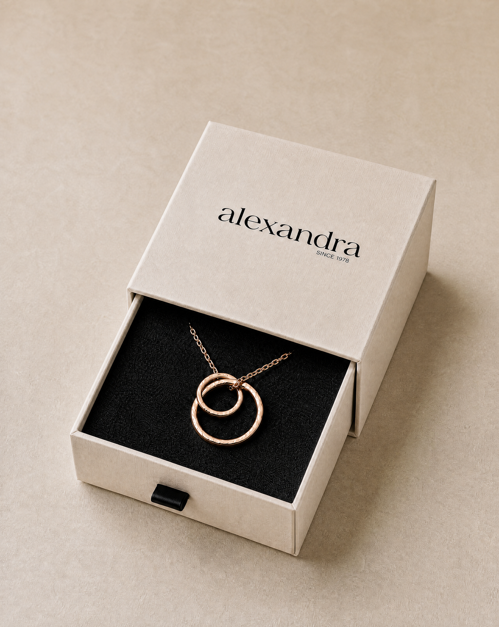 Jewelry box with a fine rose gold necklace featuring interlocking rings, branded 'alexandra', on a neutral background.