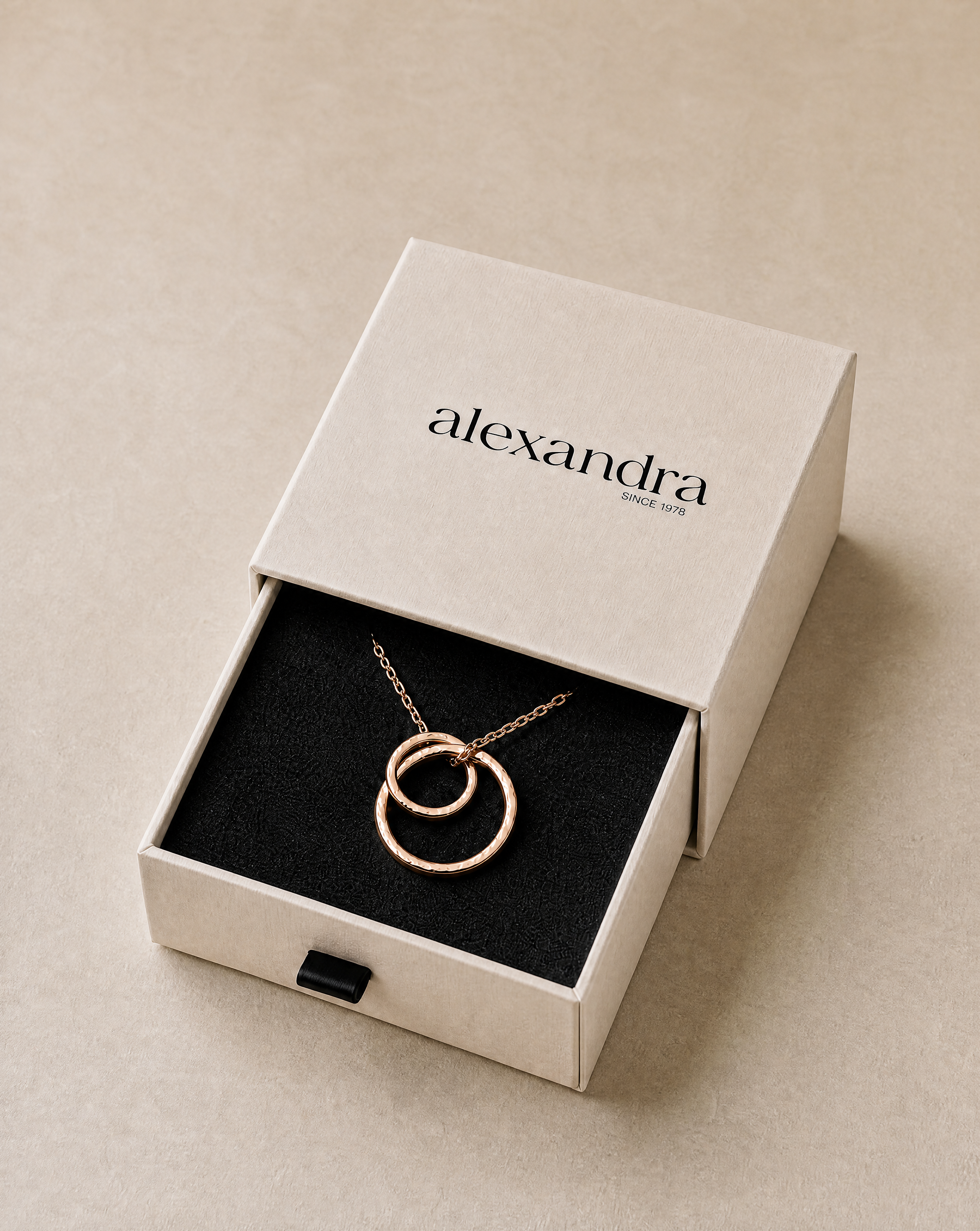 Jewelry box with a fine rose gold necklace featuring interlocking rings, branded 'alexandra', on a neutral background.