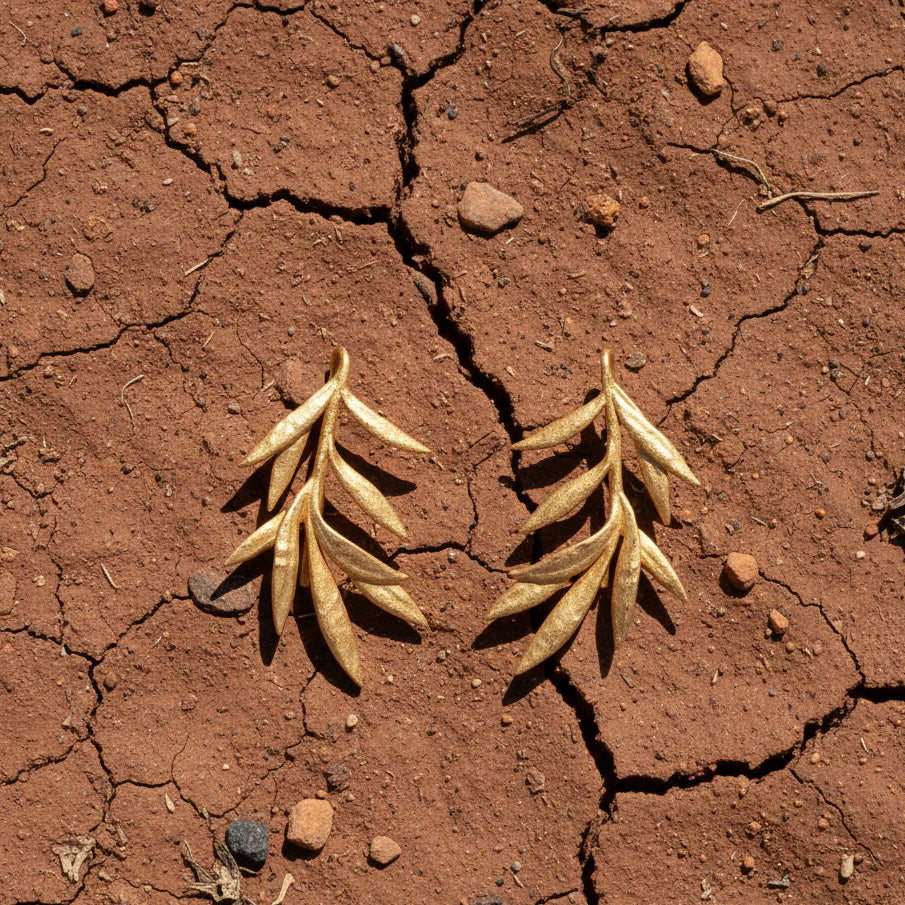 Sculptured Olive branch shaped gold stud earrings displayed on cracked, dry soil